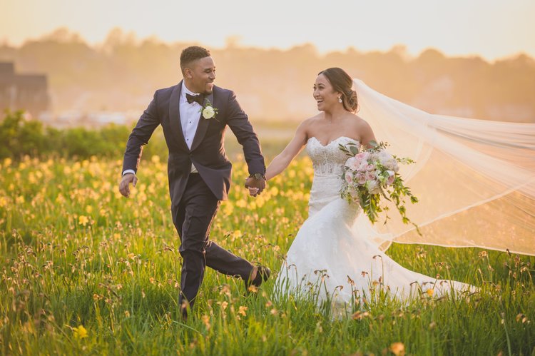 Ways To Keep Guests Cool At Your Warm-Weather Wedding - Bride And Groom Holding Hands While Running Through A Field Of Flowers.