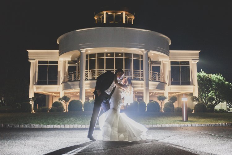 Ways To Keep Guests Cool At Your Warm-Weather Wedding - Evening Photograph Of A Bride And Groom In Front Of Their Venue With A Spotlight Shining On Them As The Groom Dips The Bride For A Kiss.