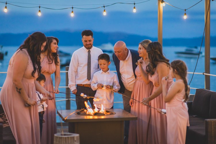 Last Minute Checklist: Things To Know Before Your Outdoor Wedding - The Bridal Party Roasting Marshmallows At An Outdoor Fire Pit During An Evening Wedding Reception.