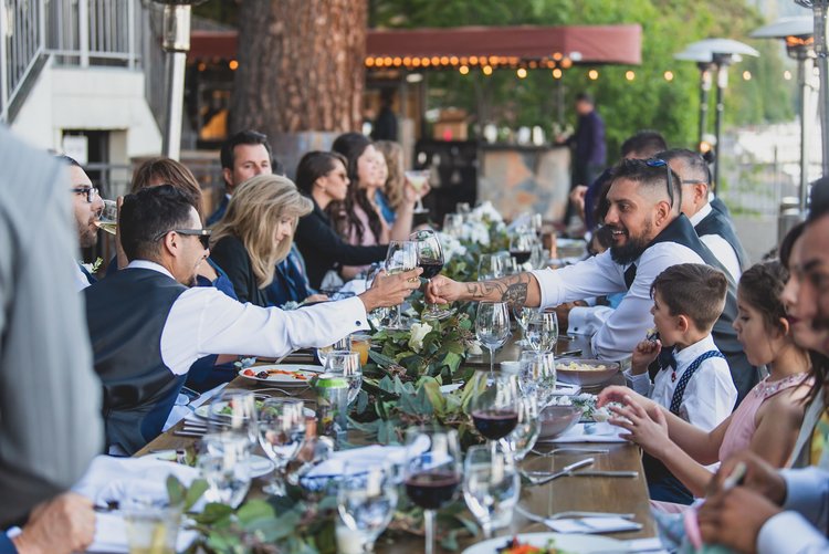 Wedding Guests Celebrate At An Outdoor Reception Sitting At A Long Wooden Table.