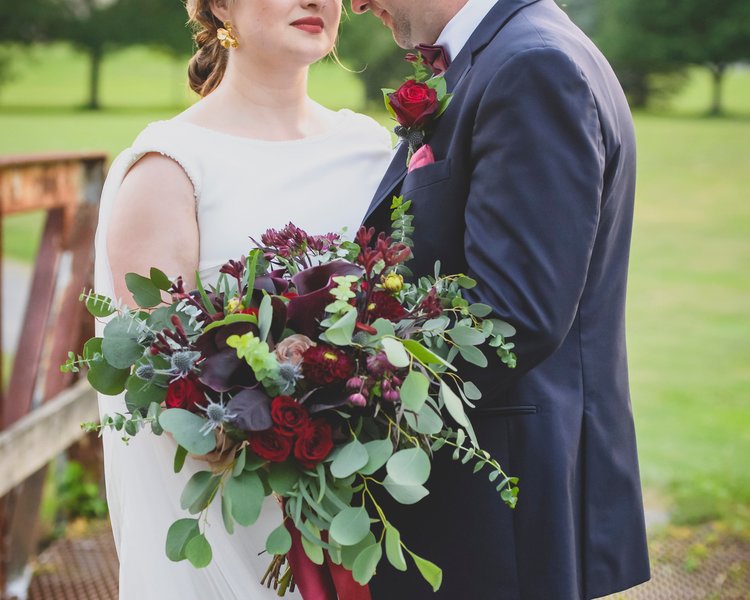 Bride And Groom Holding A Seasonal Fall Bouquet With Natural Greens And Deep Red Roses.