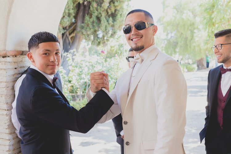 A Stylish Groom In Cream Colored Tuxedo, High-Fiving His Best Man.