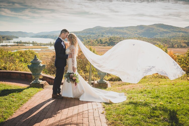 A Groom And Bride With Her Veil Blowing In The Wind Behind Them On An Outdoor Path In Front Of A Gorgeous Outdoor Backdrop Of Rivers And Rolling Hills.