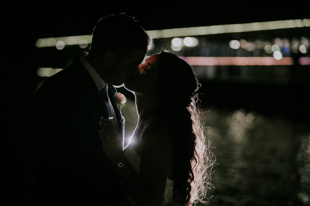 bride and groom kiss at night on waterfront while backlit by off camera flash and with tons of bokeh behind them broad ripple