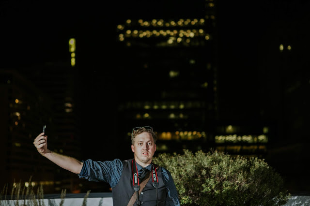 A Man Stands on Top of a Building at Night holding his phone out to provide a light source on his face