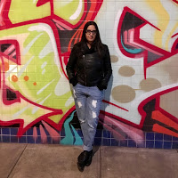 Lilliam Rivera in jeans and black leather jacket, standing before a graffiti mural wall