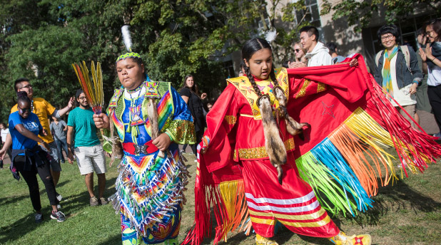 two Indigenous women in traditional regalia surrounded by TMU community in the quad