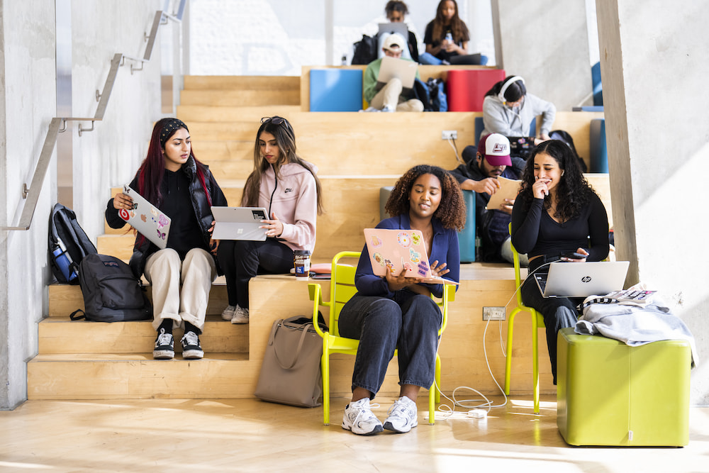 a group of students sitting on wooden stairs and brightly coloured chairs with laptops out