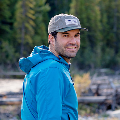 Yannis looking over his shoulder smiling standing in front of trees and a river, while wearing a baseball cap.